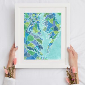 Op de afbeelding: Een ingelijst aquarel schilderij van een kaart van het Chesapeake Bay gebied. De kaart is gemaakt van veel kleine, kleurrijke vierkanten en rechthoeken. Het water is lichtblauw en het land is in verschillende tinten groen en blauw.