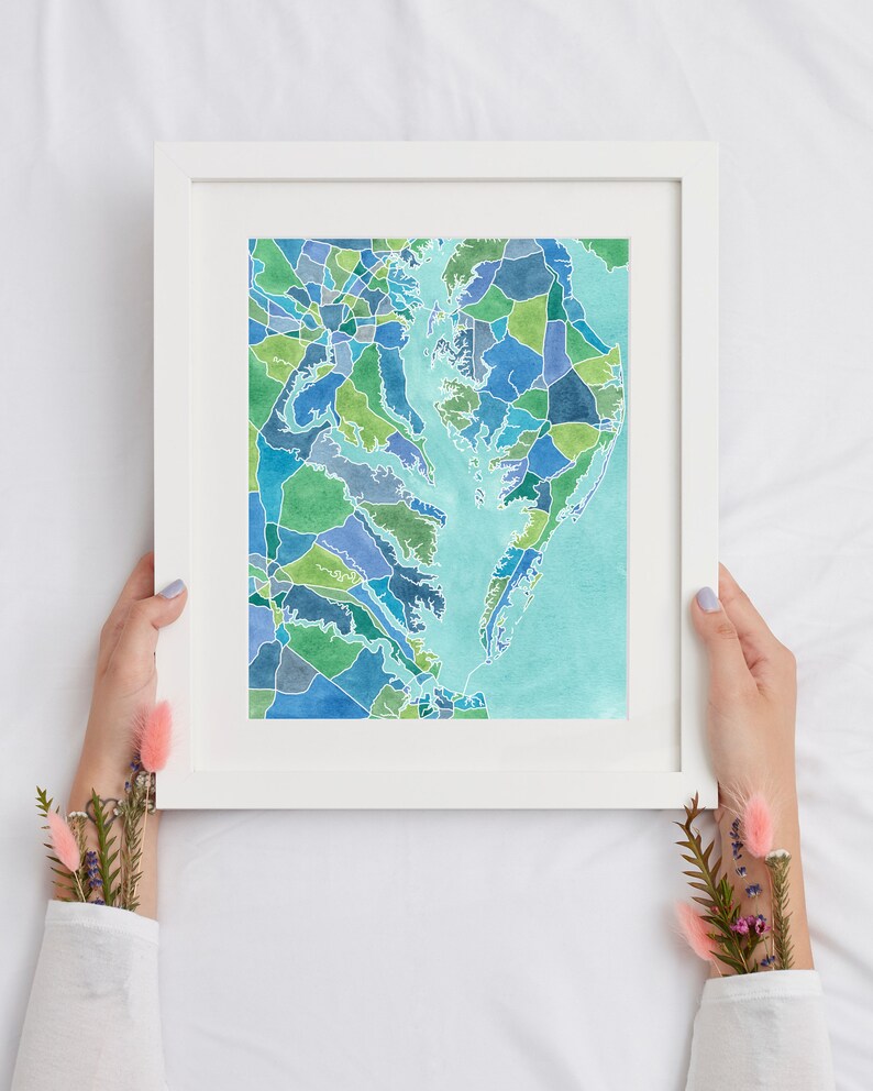 Op de afbeelding: Een ingelijst aquarel schilderij van een kaart van het Chesapeake Bay gebied. De kaart is gemaakt van veel kleine, kleurrijke vierkanten en rechthoeken. Het water is lichtblauw en het land is in verschillende tinten groen en blauw.