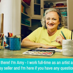 May include: A woman sits at a table with art supplies, including colored pencils, markers, and watercolors. She is smiling and holding a paintbrush. The text on the image reads "Hi there! I'm Amy - I work full-time as an artist and Etsy seller and I'm here if you have any questions!"