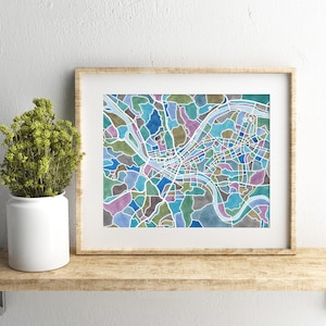 May include: A framed print of a colorful map of a city. The map is made up of different colored shapes and lines, and the river is highlighted in blue. The frame is a light brown wood.