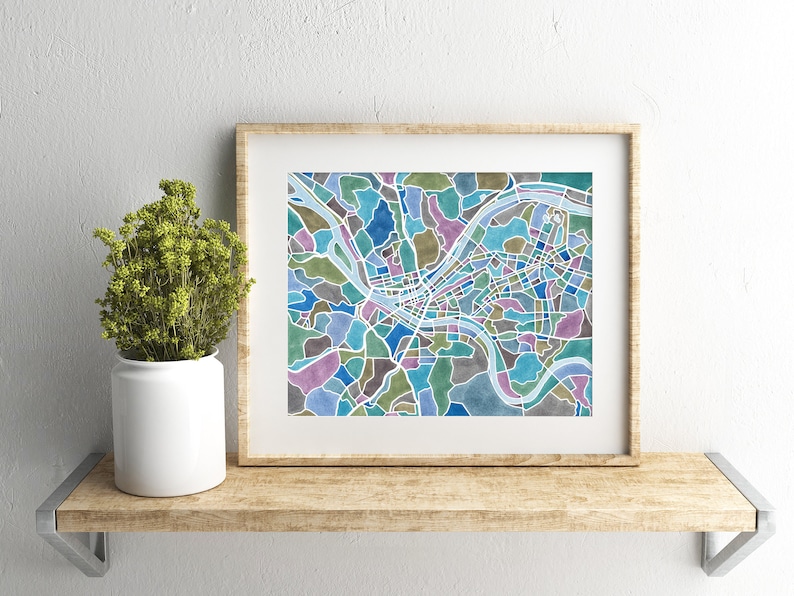 May include: A framed print of a colorful map of a city. The map is made up of different colored shapes and lines, and the river is highlighted in blue. The frame is a light brown wood.