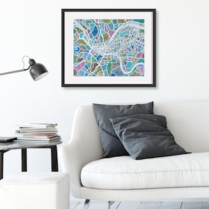 May include: A framed print of a city map with a watercolor style design. The map is in shades of blue, green, and brown. The print is hanging on a white wall above a white couch with gray pillows.