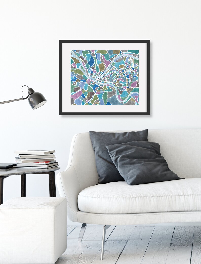 May include: A framed print of a city map with a watercolor style design. The map is in shades of blue, green, and brown. The print is hanging on a white wall above a white couch with gray pillows.