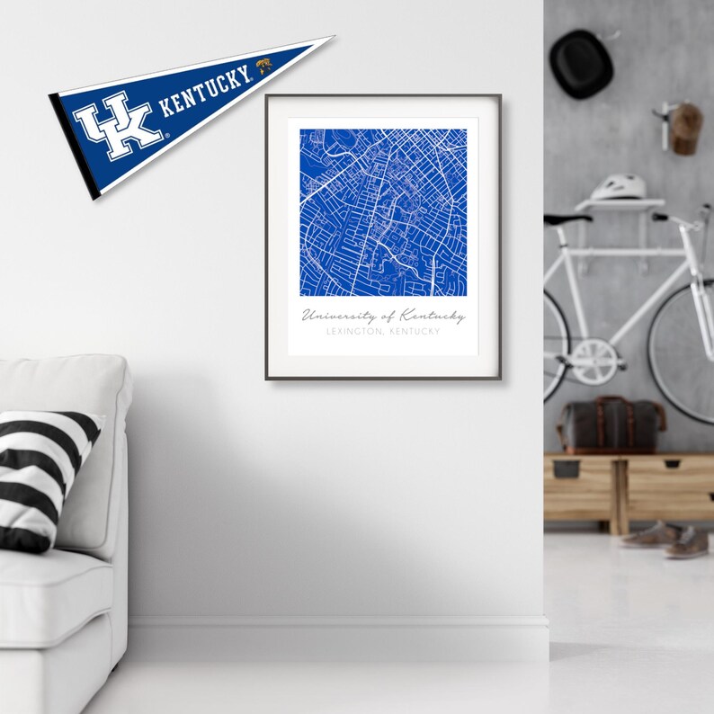 May include: A framed print of a blue map of the University of Kentucky campus in Lexington, Kentucky. The map is surrounded by a white border and a black frame. The print is hanging on a white wall.