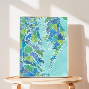 Op de afbeelding: Een ingelijst aquarel schilderij van een kaart van het Chesapeake Bay gebied. De kaart is verdeeld in kleurrijke geometrische vormen in tinten blauw, groen en turquoise.