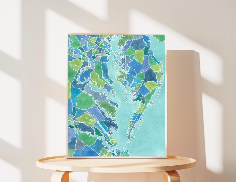 Op de afbeelding: Een ingelijst aquarel schilderij van een kaart van het Chesapeake Bay gebied. De kaart is verdeeld in kleurrijke geometrische vormen in tinten blauw, groen en turquoise.