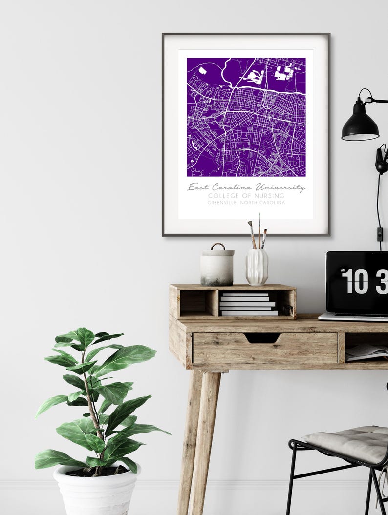 May include: A purple and white map print of East Carolina University College of Nursing in Greenville, North Carolina. The print is framed in a black frame and is hanging on a white wall.