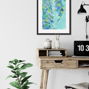 Op de afbeelding: Een ingelijst aquarel schilderij van een kaart van het Chesapeake Bay gebied, met een lichtblauwe achtergrond en een moza&iuml;ek van groene, blauwe en paarse kleuren. Het schilderij hangt aan een witte muur boven een houten bureau met een lade en een plant.