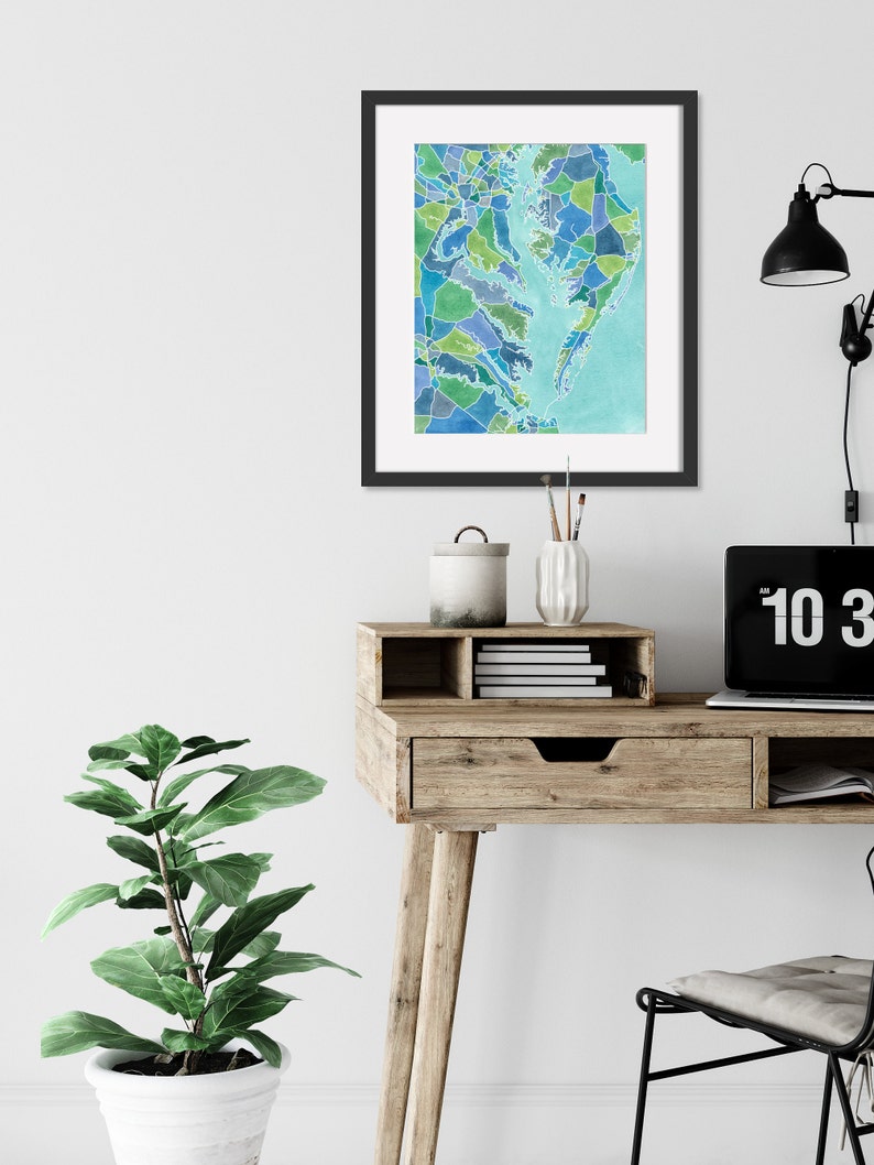 Op de afbeelding: Een ingelijst aquarel schilderij van een kaart van het Chesapeake Bay gebied, met een lichtblauwe achtergrond en een moza&iuml;ek van groene, blauwe en paarse kleuren. Het schilderij hangt aan een witte muur boven een houten bureau met een lade en een plant.