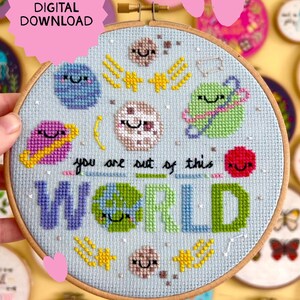 May include: A cross-stitch pattern in a wooden embroidery hoop on light blue fabric featuring colorful planets, stars, and the text "you are out of this WORLD" with a pink "DIGITAL DOWNLOAD" label in the corner of the image.