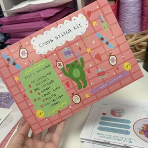 K&ouml;nnte beinhalten: Eine rosafarbene, rechteckige Schachtel f&uuml;r ein Kreuzstich-Set mit Gittermuster, einer gr&uuml;nen Frosch-Illustration und verschiedenen N&auml;hsymbolen. Die Schachtel enth&auml;lt den Text Cross stitch kit und What's included, mit der Auflistung A4 coloured pattern, DMC threads, hoop, 14 count Aida, needle und instruction sheet.