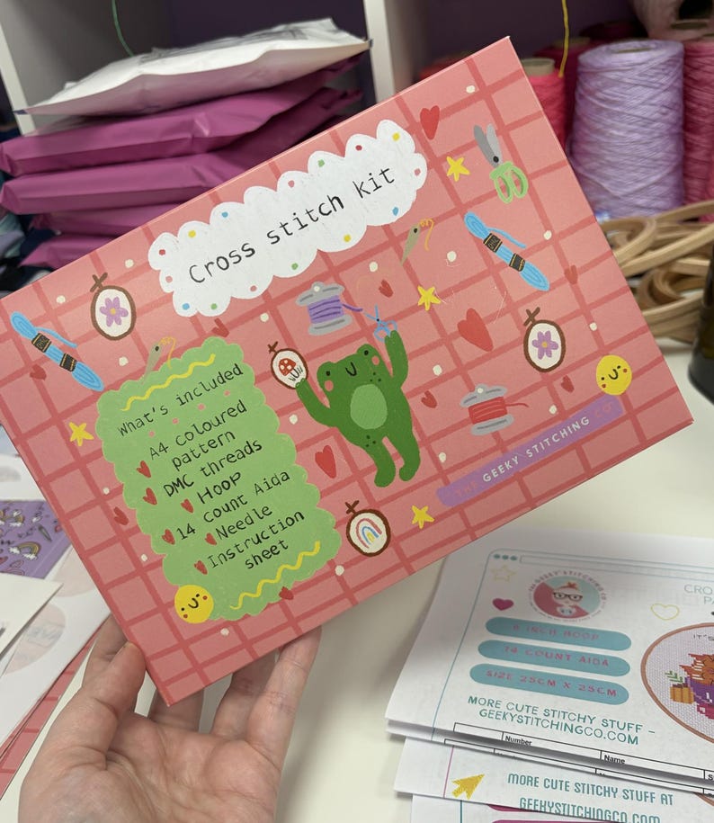 K&ouml;nnte beinhalten: Eine rosafarbene, rechteckige Schachtel f&uuml;r ein Kreuzstich-Set mit Gittermuster, einer gr&uuml;nen Frosch-Illustration und verschiedenen N&auml;hsymbolen. Die Schachtel enth&auml;lt den Text Cross stitch kit und What's included, mit der Auflistung A4 coloured pattern, DMC threads, hoop, 14 count Aida, needle und instruction sheet.
