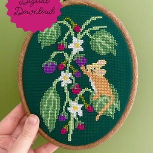 May include: Cross-stitch embroidery in a wooden hoop featuring a green background with a design of a rabbit, berries, and leaves. The text "Digital Download" is in a pink circle in the upper left corner.
