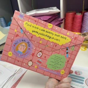 May include: A pink envelope-style folder featuring a grid pattern, decorated with illustrations of embroidery hoops, thread, and stars. It includes a yellow label with the text 'Find more cute stitchy stuff here... geekystitchingco.com' and a green label.