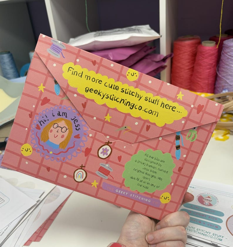 May include: A pink envelope-style folder featuring a grid pattern, decorated with illustrations of embroidery hoops, thread, and stars. It includes a yellow label with the text 'Find more cute stitchy stuff here... geekystitchingco.com' and a green label.