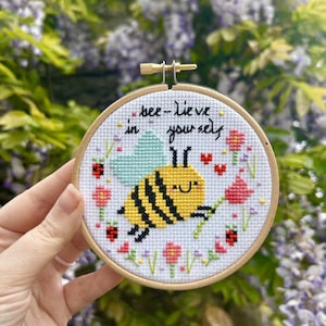 Op de afbeelding: Een ronde houten borduurring met witte stof en een kruissteekontwerp van een geel-zwarte bij met lichtblauwe vleugels. De tekst "bee-lieve in yourself" is in het zwart boven de bij geborduurd, omringd door kleine rode hartjes, roze bloemen en lieveheersbeestjes.