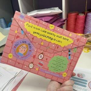 K&ouml;nnte beinhalten: Eine rosafarbene Umschlagmappe mit Karomuster, verziert mit Illustrationen von Stickrahmen, Garn und Sternen. Sie enth&auml;lt ein gelbes Etikett mit dem Text 'Find more cute stitchy stuff here... geekystitchingco.com' sowie ein gr&uuml;nes Etikett.