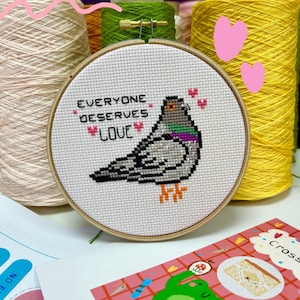 K&ouml;nnte beinhalten: Ein runder Holzstickrahmen mit wei&szlig;em Stoff, der ein Kreuzstichmotiv einer grauen Taube mit violettem und gr&uuml;nem Hals zeigt. Der Text lautet EVERYONE DESERVES LOVE, erg&auml;nzt durch kleine, um den Vogel herum gestickte rosa Herzen.