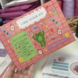 May include: A pink rectangular cross stitch kit box featuring a grid pattern, a green frog illustration, and various sewing icons. The box includes text reading Cross stitch kit and What's included, listing A4 coloured pattern, DMC threads, hoop, 14 count Aida, needle, and instruction sheet.