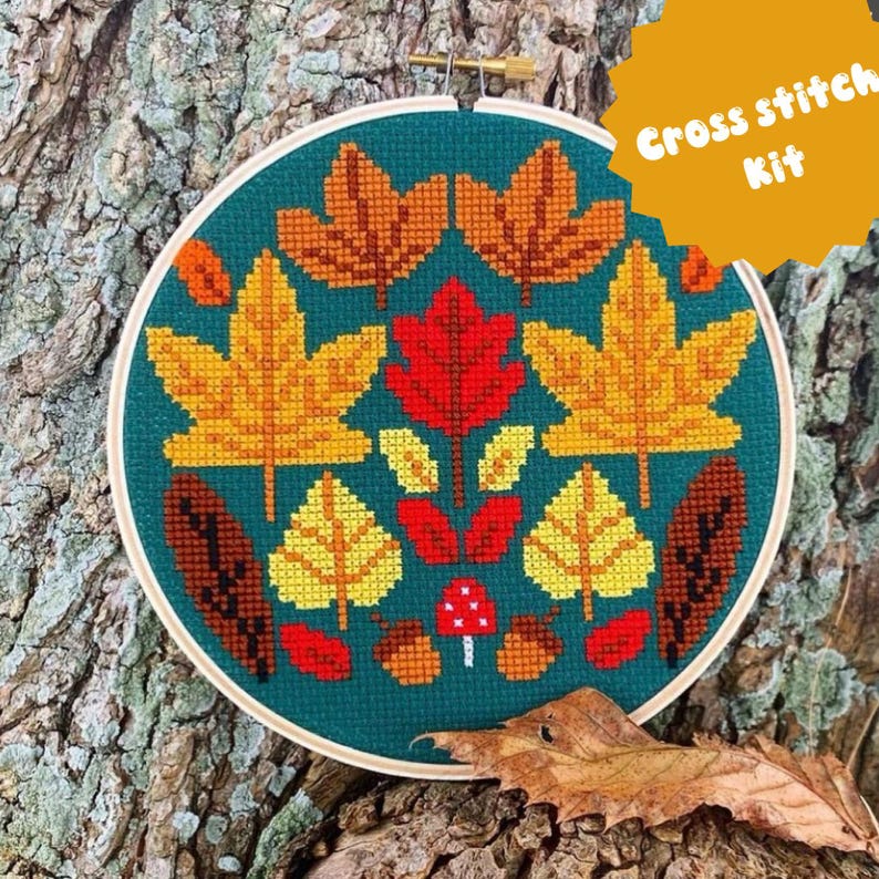 May include: A circular wooden embroidery hoop containing a cross stitch pattern on dark teal fabric. The design features various autumn leaves in yellow, orange, and red, along with brown acorns and a small red mushroom with white spots.