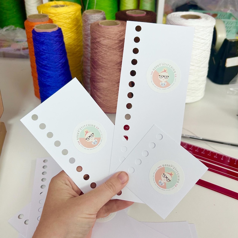 May include: Three white paper thread organisers with circular holes along one edge and a circular logo sticker featuring a cartoon character with glasses and the text The Geeky Stitching Co. arranged in front of colourful spools of yarn.