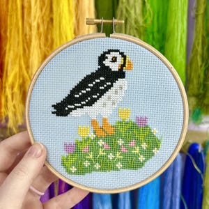 Könnte beinhalten: Eine Kreuzstichstickerei in einem runden Holzrahmen, die einen Papageientaucher mit schwarz-weißem Gefieder und orangefarbenem Schnabel zeigt. Der Vogel steht auf einer grünen Grasfläche, die mit kleinen gelben, rosa und violetten Blumen auf hellblauem Grund verziert ist.