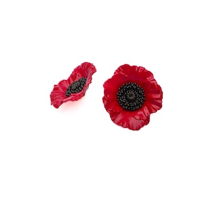 May include: Two poppy flower ornaments featuring vibrant red, ruffled petals and a textured, dark black center. One flower is positioned flat, displaying its full circular shape, while the other is angled to show its curved, dimensional profile.