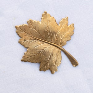 May include: A gold-colored metal leaf with detailed veins and a stem. The leaf is on a white background.