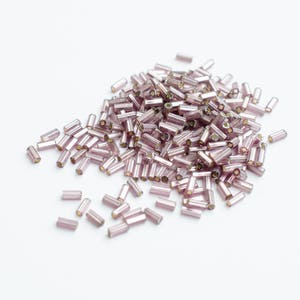 May include: A pile of small, light purple glass beads. The beads are cylindrical in shape and have a slight shimmer.