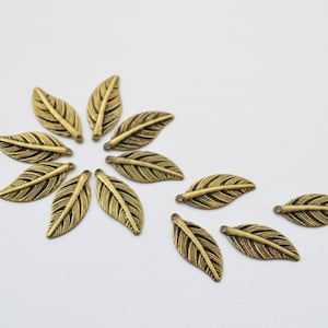 May include: A collection of gold-toned metal leaf charms with a textured surface. The charms are arranged in a circular pattern on a white background.