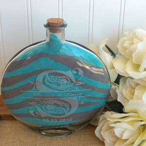 May include: A round glass bottle with a cork stopper filled with teal and gray sand. The bottle has a zebra stripe pattern and is engraved with the letter "S", the names "Monica & Larry" and the date "10.23.2016".