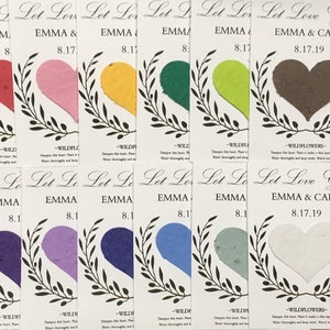 May include: A collection of seed paper wedding favors with heart-shaped wildflower seeds in various colors. Each favor features the text "Let Love Grow" and the names "EMMA & CARL" with the date "8.17.19".