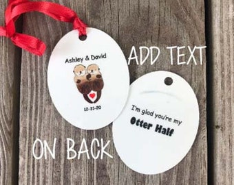 May include: Two round white ornaments with a red ribbon. The front ornament features a cartoon image of two otters holding a red heart with the text "Ashley & David 12-31-20". The back ornament has the text "I'm glad you're my Otter Half".