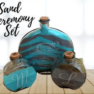 May include: Sand ceremony set with three glass bottles. The bottles are decorated with black and teal stripes and have cork tops. The bottles are labeled with the letters "M" and "L".