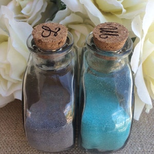 May include: Two small glass bottles with cork tops. The bottles are filled with sand, one is gray and the other is blue. The cork tops have initials carved into them, "S" and "M".