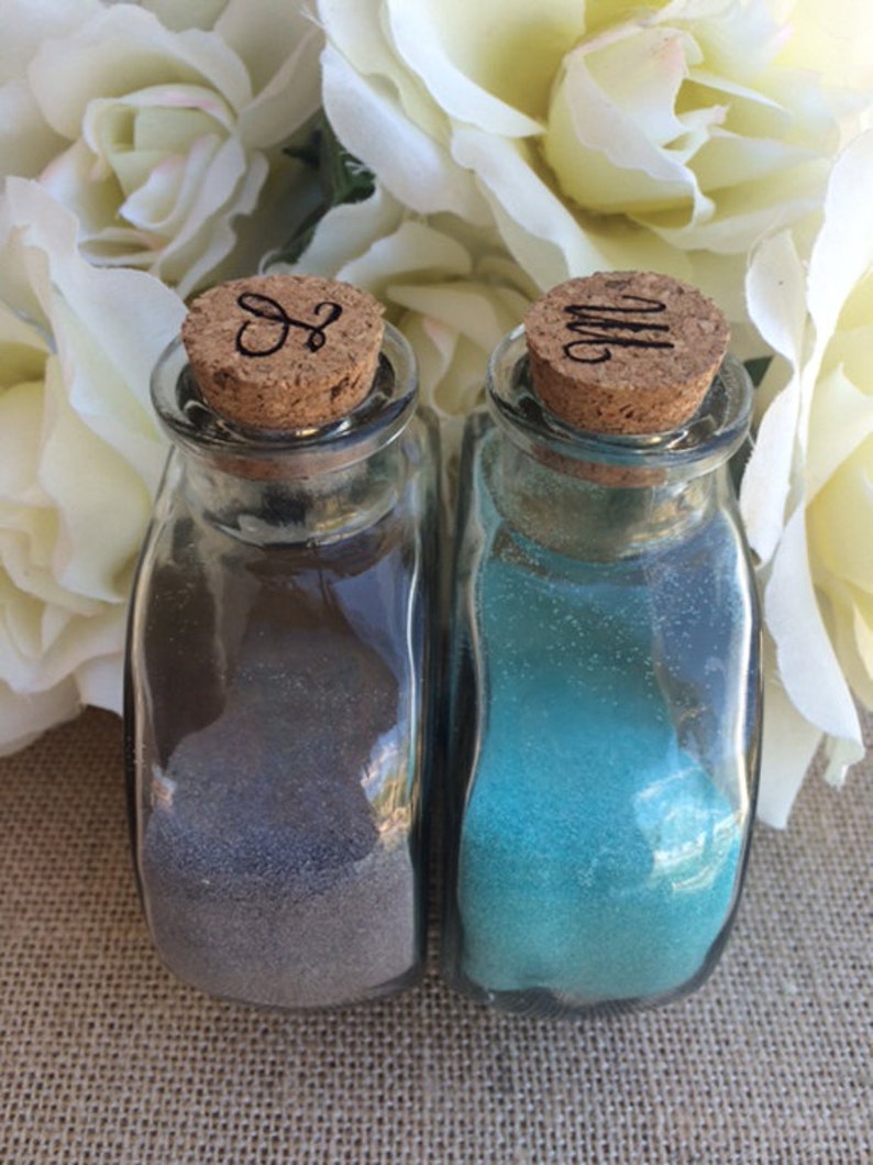 May include: Two small glass bottles with cork tops. The bottles are filled with sand, one is gray and the other is blue. The cork tops have initials carved into them, "S" and "M".