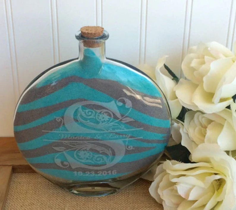 May include: A round glass bottle with a cork stopper filled with teal and gray sand. The bottle is etched with the letter "S" and the text "Monica & Larry 10.23.2016".