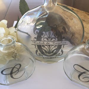May include: Three clear glass bottles with black lettering. The largest bottle has the text "Caleb & Taylor" and "5.13.2017" on it. The two smaller bottles have the letters "C" and "T" on them.