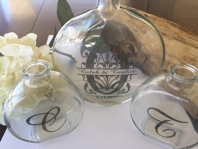 May include: Three clear glass bottles with black lettering. The largest bottle has the text "Caleb & Taylor" and "5.13.2017" on it. The two smaller bottles have the letters "C" and "T" on them.