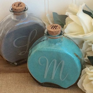 May include: Two round glass bottles with cork stoppers, filled with sand. The bottles are decorated with the letters "L" and "M" on the front. The bottles are sitting on a wooden surface.