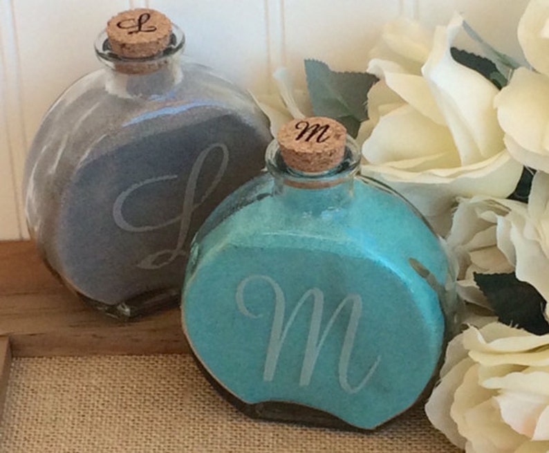 May include: Two round glass bottles with cork stoppers, filled with sand. The bottles are decorated with the letters "L" and "M" on the front. The bottles are sitting on a wooden surface.