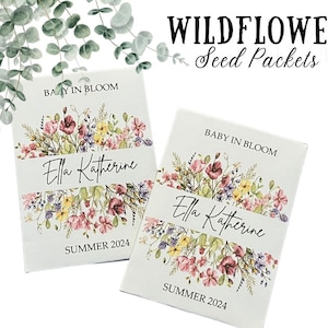May include: Two white seed packets with a floral design and the text "BABY IN BLOOM" and "Ella Katherine" with the year "SUMMER 2024" printed on them. The packets are labeled "WILDFLOWER Seed Packets".