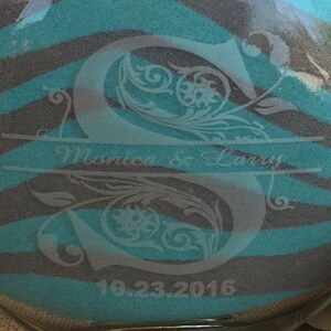 May include: A clear glass sand ceremony vase with a teal and gray zebra stripe pattern. The vase is etched with the letters "S" and the text "Monica & Larry" and the date "10.23.2016".