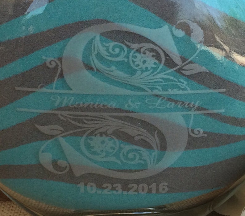 May include: A clear glass sand ceremony vase with a teal and gray zebra stripe pattern. The vase is etched with the letters "S" and the text "Monica & Larry" and the date "10.23.2016".