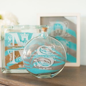 May include: Two glass sand ceremony containers with personalized engravings. The larger container is a square block with the letter "M" and the name "Christopher". The smaller container is a round bottle with the letter "S" and the names "Monica & Larry" and the date "10.23.2016".