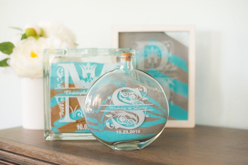 May include: Two glass sand ceremony containers with personalized engravings. The larger container is a square block with the letter "M" and the name "Christopher". The smaller container is a round bottle with the letter "S" and the names "Monica & Larry" and the date "10.23.2016".