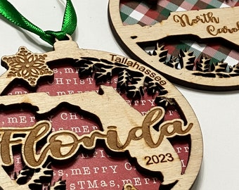 May include: Two wooden Christmas ornaments with a red and green plaid design and a snowflake. The ornaments are decorated with the words "North Carolina" and "Tallahassee" and the state of Florida. The ornament for Florida has the year 2023 on it.