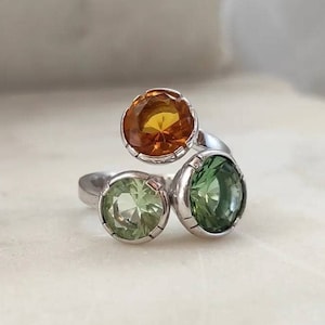 May include: A silver ring with three round gemstones. The largest gemstone is orange, and the two smaller gemstones are green.