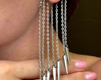 May include: Silver ear cuff with multiple dangling chains ending in sharp, pointed tips. The chains are connected to a hoop that goes through the ear.
