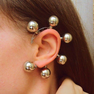May include: Silver ear cuff with multiple round balls attached. The cuff is designed to wrap around the ear and has a curved shape.